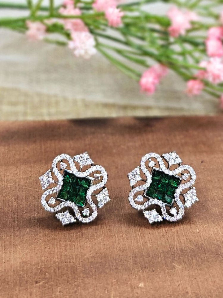 The Green Zuri Earrings feature ornate Green square gemstones at the center, accented by intricate silver designs and sparkling American diamonds, displayed on a brown surface with pink flowers in the background.