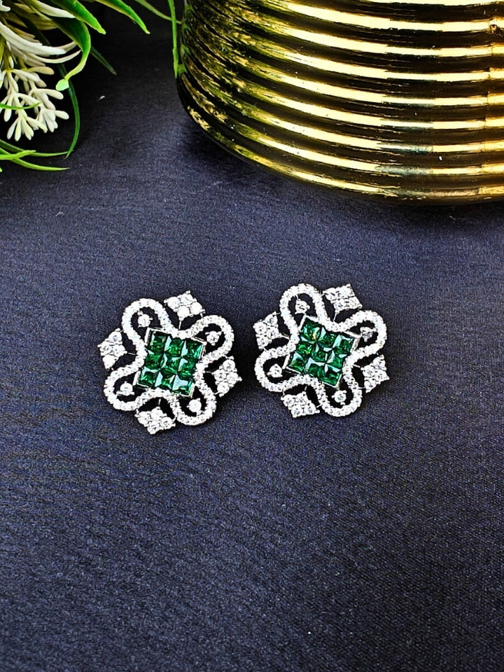 The Green Zuri Earrings feature ornate Green square gemstones at the center, accented by intricate silver designs and sparkling American diamonds, displayed on a Black surface with White flowers in the background.