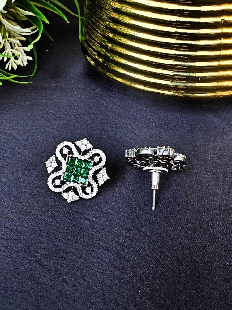 The Green Zuri Earrings feature ornate Green square gemstones at the center, accented by intricate silver designs and sparkling American diamonds, displayed on a Black surface with White flowers in the background.