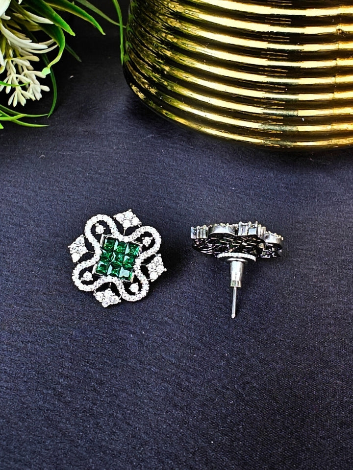 The Green Zuri Earrings feature ornate Green square gemstones at the center, accented by intricate silver designs and sparkling American diamonds, displayed on a Black surface with White flowers in the background.