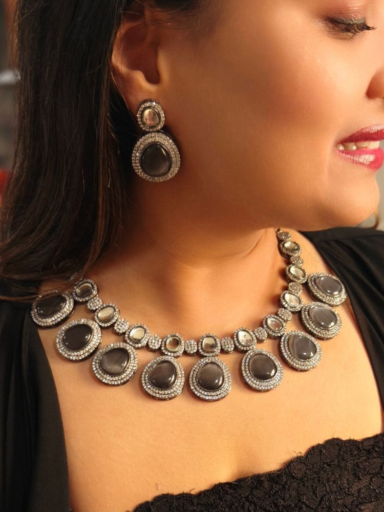 A woman with brown hair down smiles to the side, wearing the Grey Elizabeth Necklace Set featuring a statement Victorian-style necklace and matching earrings with large colorful stones, paired with a sparkly dark top.