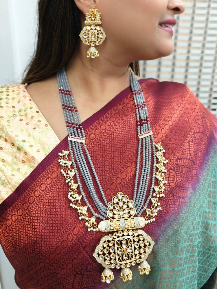 A woman in a traditional red, gold, and green silk saree wears the Grey Shazia Necklace Set with matching earrings and poses in profile with a smile—an elegant choice for a wedding ceremony. Kundan Meenakari pendant hanging with grey crystal stones, and pearls, perfect for weddings. 