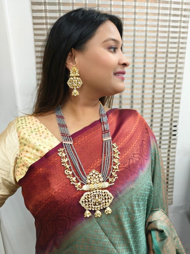 A woman in a traditional red, gold, and green silk saree wears the Grey Shazia Necklace Set with matching earrings and poses in profile with a smile—an elegant choice for a wedding ceremony. Kundan Meenakari pendant hanging with grey crystal stones, and pearls, perfect for weddings. 