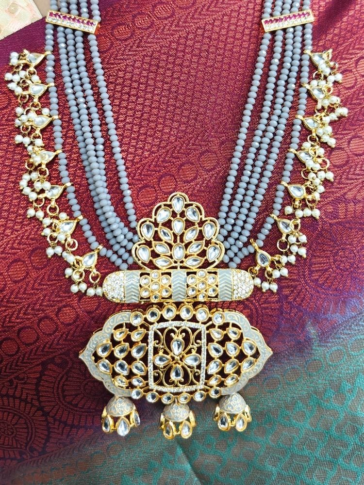 A woman in a traditional red, gold, and green silk saree wears the Grey Shazia Necklace Set with matching earrings and poses in profile with a smile—an elegant choice for a wedding ceremony. Kundan Meenakari pendant hanging with grey crystal stones, and pearls, perfect for weddings. 