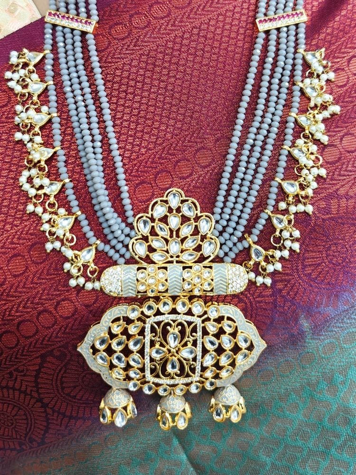 A woman in a traditional red, gold, and green silk saree wears the Grey Shazia Necklace Set with matching earrings and poses in profile with a smile—an elegant choice for a wedding ceremony. Kundan Meenakari pendant hanging with grey crystal stones, and pearls, perfect for weddings. 