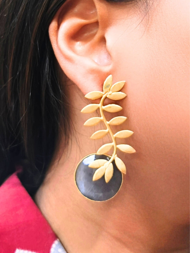 A close-up of a womans ear wearing Haley Grey Earrings—large brass with a leafy branch design and a round, translucent pale Grey stone at the bottom. Her dark hair and part of a pink patterned garment are also visible. Casual wear earrings. 
