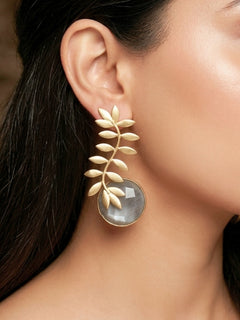 A close-up of a womans ear wearing Haley Grey Earrings—large brass with a leafy branch design and a round, translucent Grey stone at the bottom. Her dark hair and part of a patterned garment are also visible. Casual wear earrings. 