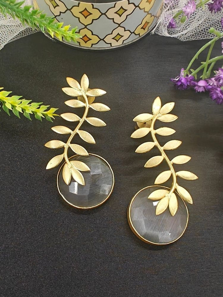 A close-up of Haley Grey Earrings—large brass with a leafy branch design and a round, translucent Grey stone at the bottom. Her dark hair and part of a patterned garment are also visible. Casual wear earrings. 