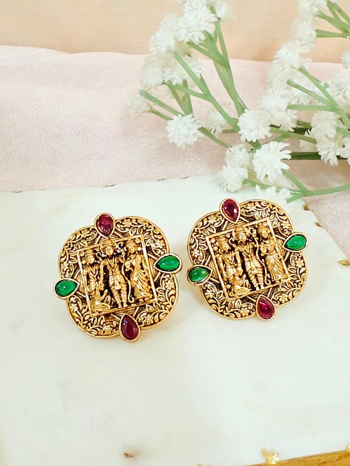 The Hari Earrings are ornate gold-toned brass earrings with Ram, Laxman and Sita placed in the middle, along and pink & green stones, displayed on a white surface with white flowers in the background.