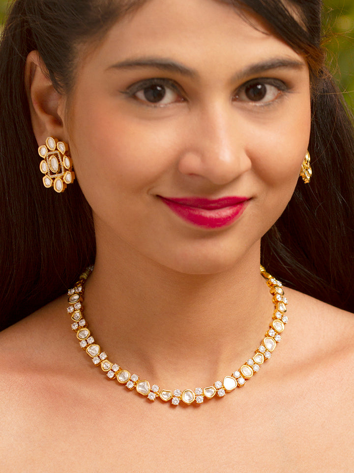 A Harley necklace set with a gold-colored chain and a pair of matching earrings, adorned with white and yellow diamonds, displayed on a dark background with a cup of tea and yellow flowers in the background.

