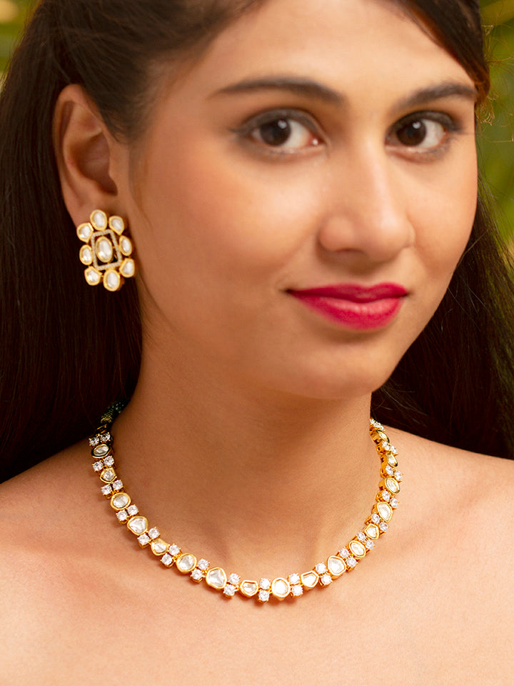 A Harley necklace set with a gold-colored chain and a pair of matching earrings, adorned with white and yellow diamonds, displayed on a dark background with a cup of tea and yellow flowers in the background.

