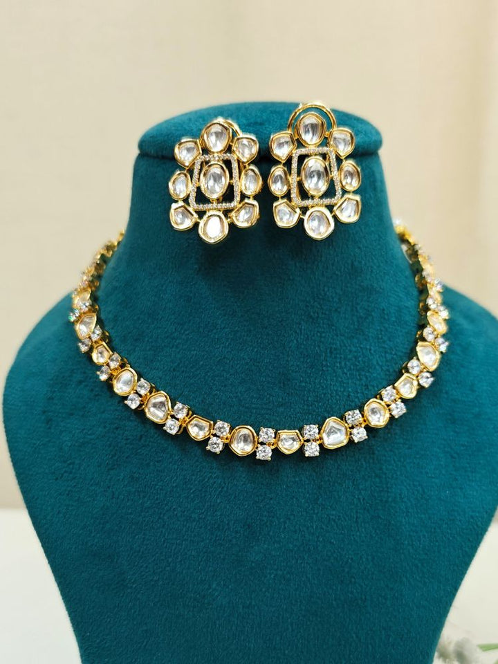 A Harley necklace set with a gold-colored chain and a pair of matching earrings, adorned with white and yellow diamonds, displayed on a dark background with a cup of tea and yellow flowers in the background.

