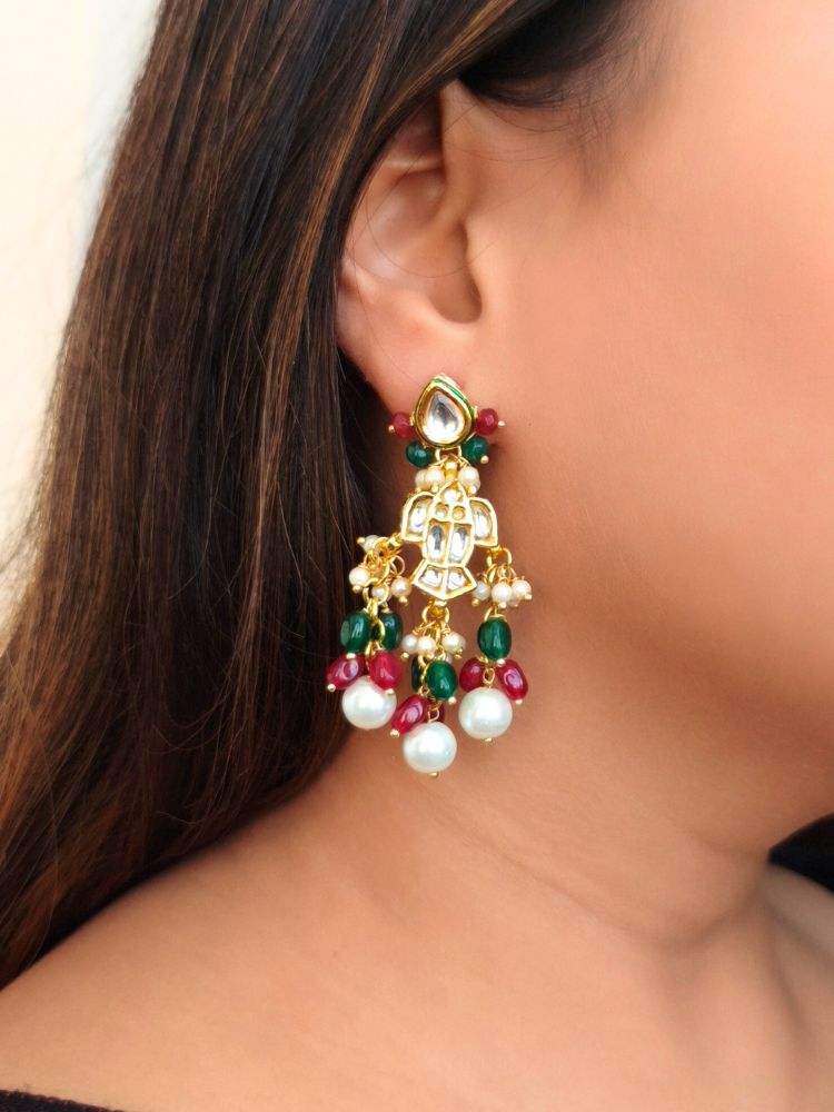 A close-up shot shows a womans ear wearing Harper Earrings—large Kundan gold pieces with red and green beads and dangling pearls, ideal for pairing with a wedding ceremony dress. She has long, straight hair and wears a dark top.