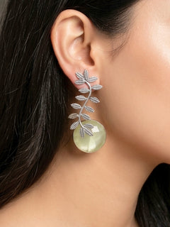 A close-up of a womans ear wearing Harriet Lemon Yellow Earrings—large brass with a leafy branch design and a round, translucent Lemon yellow stone at the bottom. Her dark hair and part of a pink patterned garment are also visible. Casual wear earrings.  Festive celebration look enhanced with artificial Oxidised Earrings.