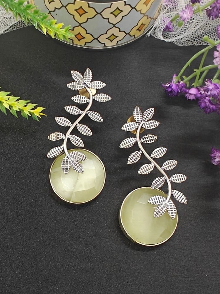 The Harriet Lemon Yellow Earrings feature oxidized polish, leaf-shaped designs on top, and large a large yellow round stone at the bottom, displayed on black fabric with green and purple flowers and a patterned ceramic pot in the background.