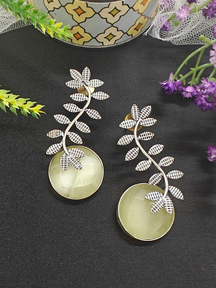 The Harriet Lemon Yellow Earrings feature oxidized polish, leaf-shaped designs on top, and large a large yellow round stone at the bottom, displayed on black fabric with green and purple flowers and a patterned ceramic pot in the background.