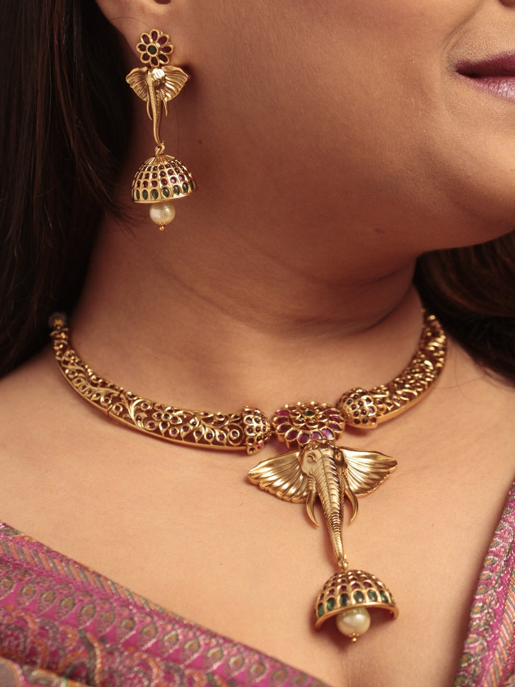 A woman wearing the Hastika Hasli Necklace Set featuring a traditional gold Collar hasli necklace and matching earrings with Goddess Ganesh in the middle, along with pink and green colored stones and golden elephants, paired with a Saree—ideal party jewelry for any festive occasion. Festive Diwali look completed with a traditional temple necklace set.