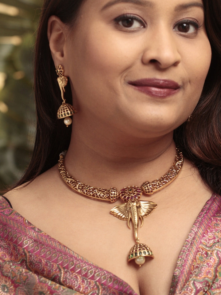 A woman wearing the Hastika Hasli Necklace Set featuring a traditional gold Collar hasli necklace and matching earrings with Goddess Ganesh in the middle, along with pink and green colored stones and golden elephants, paired with a Saree—ideal party jewelry for any festive occasion. Festive Diwali look completed with a traditional temple necklace set.