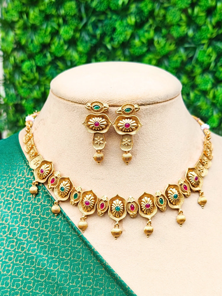 The Heena Necklace Set, featuring gold floral designs with ruby and emerald accents is perfect for south indian jewellery, is displayed on a mannequin draped in green patterned fabric against a leafy background.