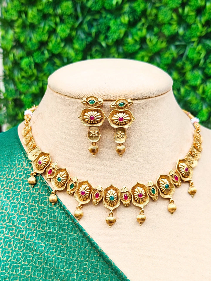 The Heena Necklace Set, featuring gold floral designs with ruby and emerald accents is perfect for south indian jewellery, is displayed on a mannequin draped in green patterned fabric against a leafy background.