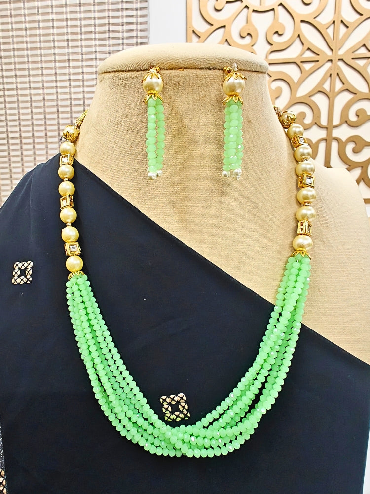The Hiran Mint Green Necklace Set is displayed on a black-draped mannequin, featuring green beaded multi-layer strands, crystal stones, gold accents, and matching green beaded drop earrings against a decorative background.