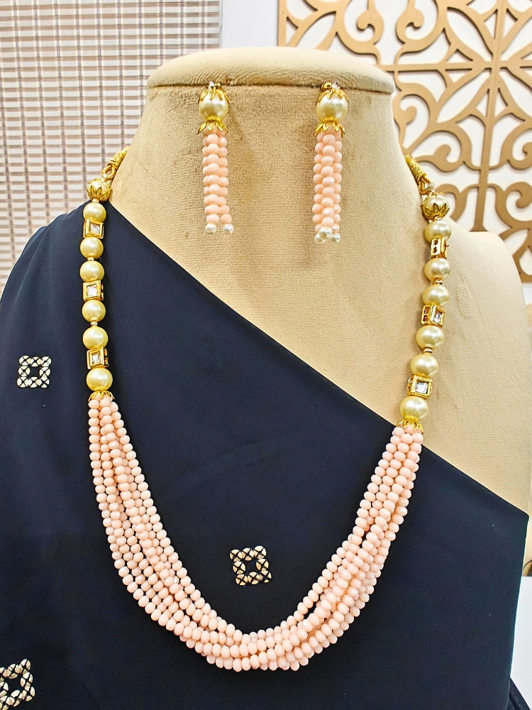 The Hiran Mint Peach Necklace Set is displayed on a black-draped mannequin, featuring Peach beaded multi-layer strands, crystal stones, gold accents, and matching Peach beaded drop earrings against a decorative background.