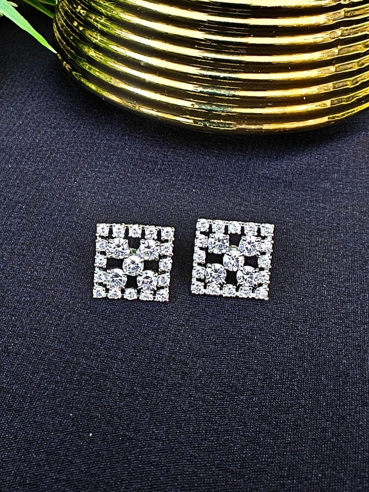 The Hope White Earrings are square studs with central White stones in a cross pattern, surrounded by sparkling American diamonds, shown on a dark surface with gold-ridged decor and green leaves.