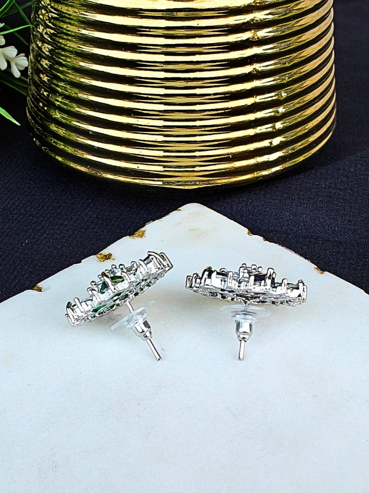 The Hope White Earrings are square studs with central White stones in a cross pattern, surrounded by sparkling American diamonds, shown on a White Marble surface with gold-ridged decor and green leaves.