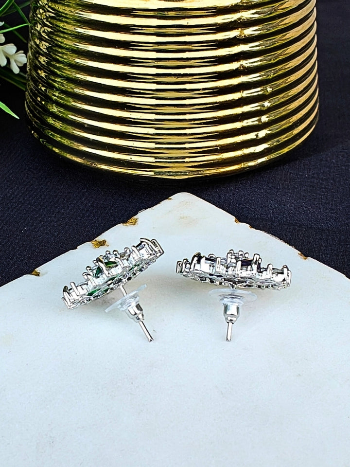 The Hope White Earrings are square studs with central White stones in a cross pattern, surrounded by sparkling American diamonds, shown on a White Marble surface with gold-ridged decor and green leaves.