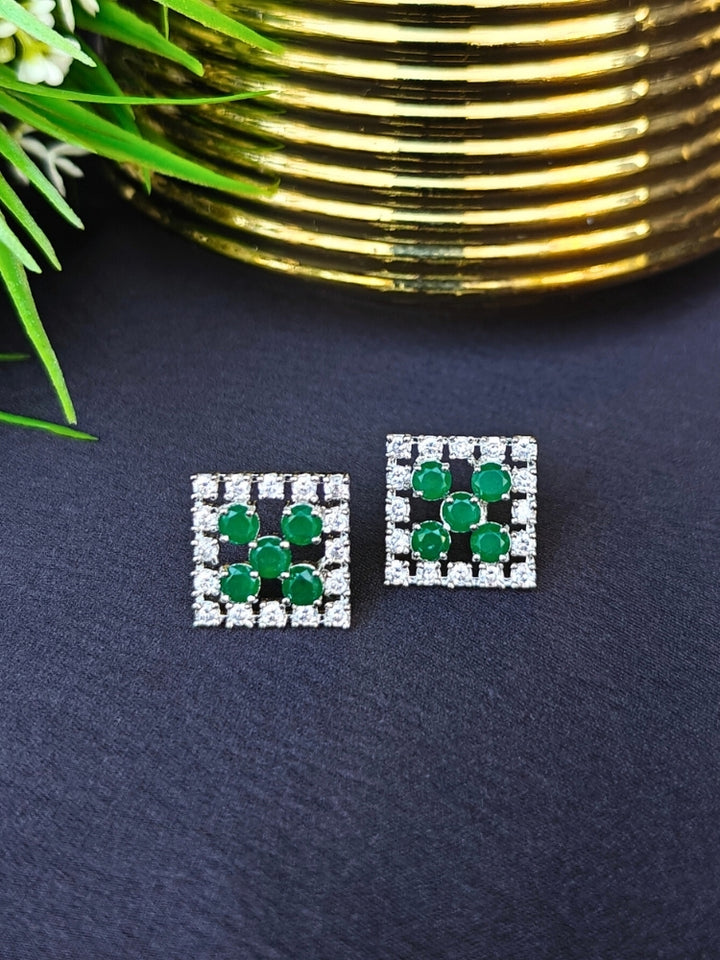 The Hope Green Earrings are square studs with central emerald green stones in a cross pattern, surrounded by sparkling American diamonds, shown on a dark surface with gold-ridged decor and green leaves.