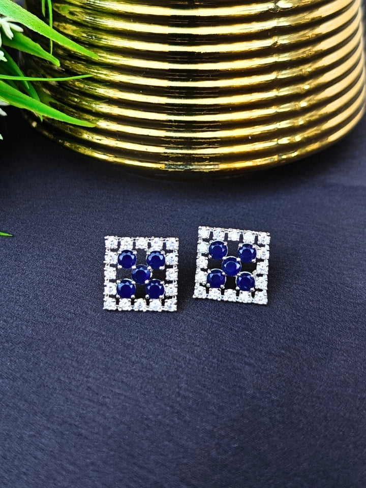 The Hope Navy Blue Earrings are square studs with central Navy Blue stones in a cross pattern, surrounded by sparkling American diamonds, shown on a dark surface with gold-ridged decor and green leaves.