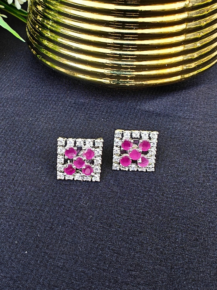The Hope Pink Earrings are square studs with central Pink stones in a cross pattern, surrounded by sparkling American diamonds, shown on a dark surface with gold-ridged decor and green leaves.