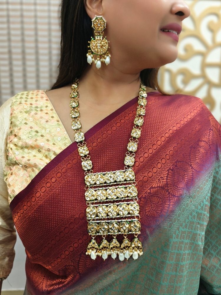 A woman in a red and green saree smiles softly, wearing the exquisite Ikshita Necklace Set made entired with kundan along with pearls hanging at the bottom of the pendant and earrings, posing against a patterned background.