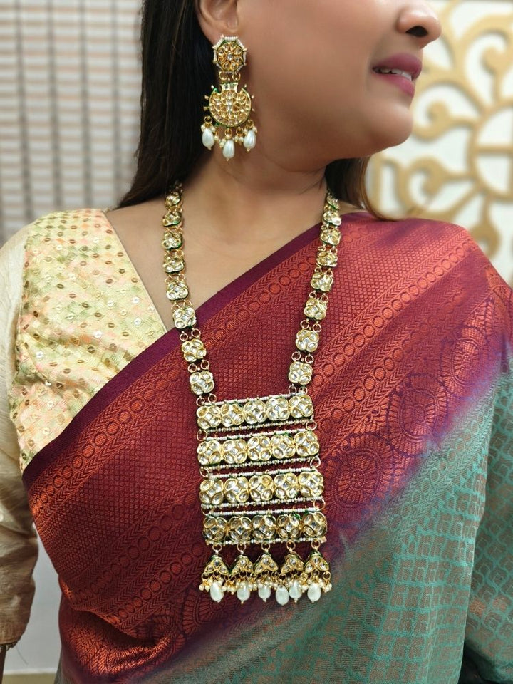 A woman in a red and green saree smiles softly, wearing the exquisite Ikshita Necklace Set made entired with kundan along with pearls hanging at the bottom of the pendant and earrings, posing against a patterned background.