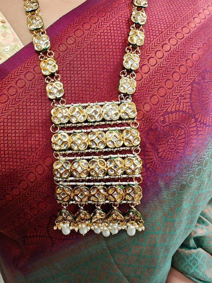 A woman in a red and green saree smiles softly, wearing the exquisite Ikshita Necklace Set made entired with kundan along with pearls hanging at the bottom of the pendant and earrings, posing against a patterned background.