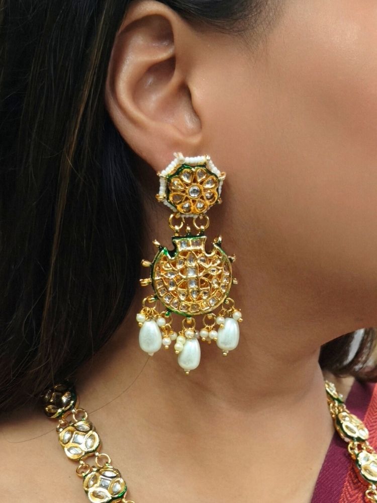 A woman in a red and green saree smiles softly, wearing the exquisite Ikshita Necklace Set made entired with kundan along with pearls hanging at the bottom of the pendant and earrings, posing against a patterned background.