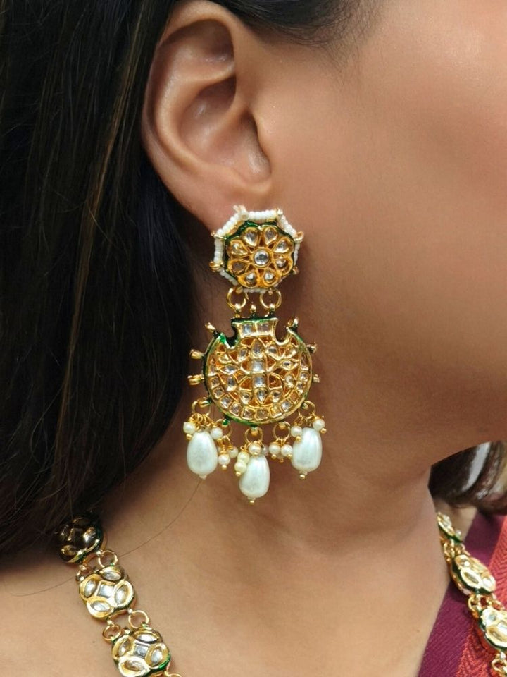 A woman in a red and green saree smiles softly, wearing the exquisite Ikshita Necklace Set made entired with kundan along with pearls hanging at the bottom of the pendant and earrings, posing against a patterned background.