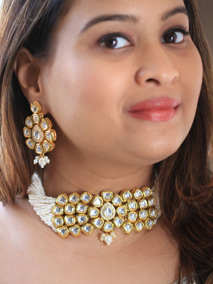 A woman smiles softly, her hair down and makeup natural with a rosy lip, as she wears the Ishika Choker Necklace Set with matching statement earrings. Made with multiple strands of pearls, with a big kundan center piece along with matching kundan long earrings, this is a perfect wedding necklace. 