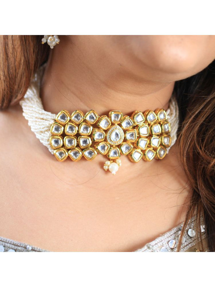 A woman smiles softly, her hair down and makeup natural with a rosy lip, as she wears the Ishika Choker Necklace Set with matching statement earrings. Made with multiple strands of pearls, with a big kundan center piece along with matching kundan long earrings, this is a perfect wedding necklace. 