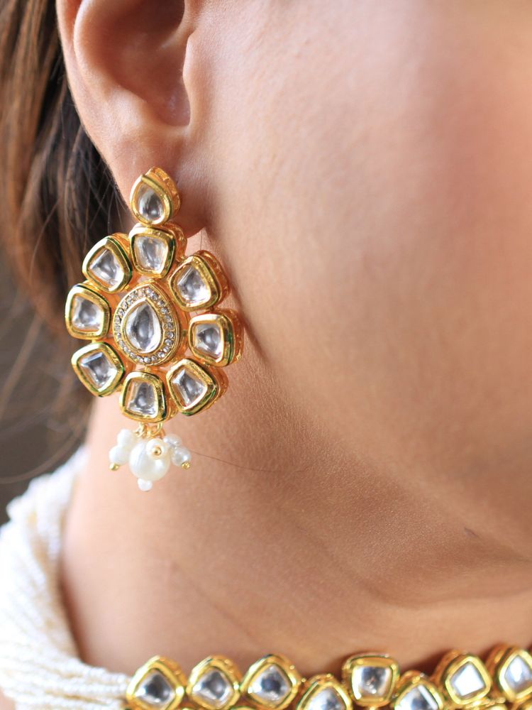 A woman smiles softly, her hair down and makeup natural with a rosy lip, as she wears the Ishika Choker Necklace Set with matching statement earrings. Made with multiple strands of pearls, with a big kundan center piece along with matching kundan long earrings, this is a perfect wedding necklace. 