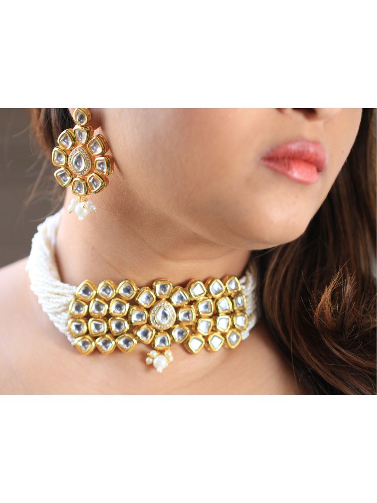 A woman smiles softly, her hair down and makeup natural with a rosy lip, as she wears the Ishika Choker Necklace Set with matching statement earrings. Made with multiple strands of pearls, with a big kundan center piece along with matching kundan long earrings, this is a perfect wedding necklace. 