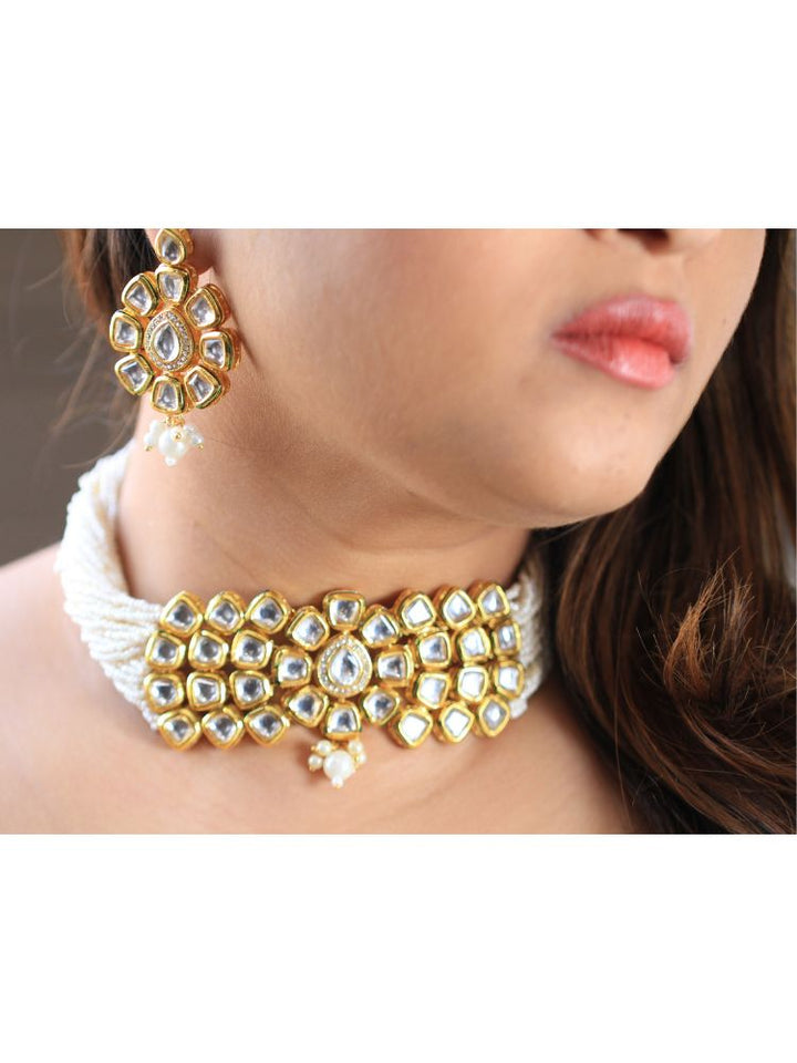 A woman smiles softly, her hair down and makeup natural with a rosy lip, as she wears the Ishika Choker Necklace Set with matching statement earrings. Made with multiple strands of pearls, with a big kundan center piece along with matching kundan long earrings, this is a perfect wedding necklace. 