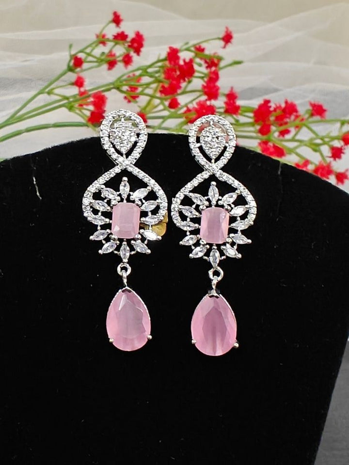 The Ivanka Earrings are elegant pink gemstone drop earrings, adorned with sparkling American Diamonds in intricate silver settings, showcased on a black background with red flowers.