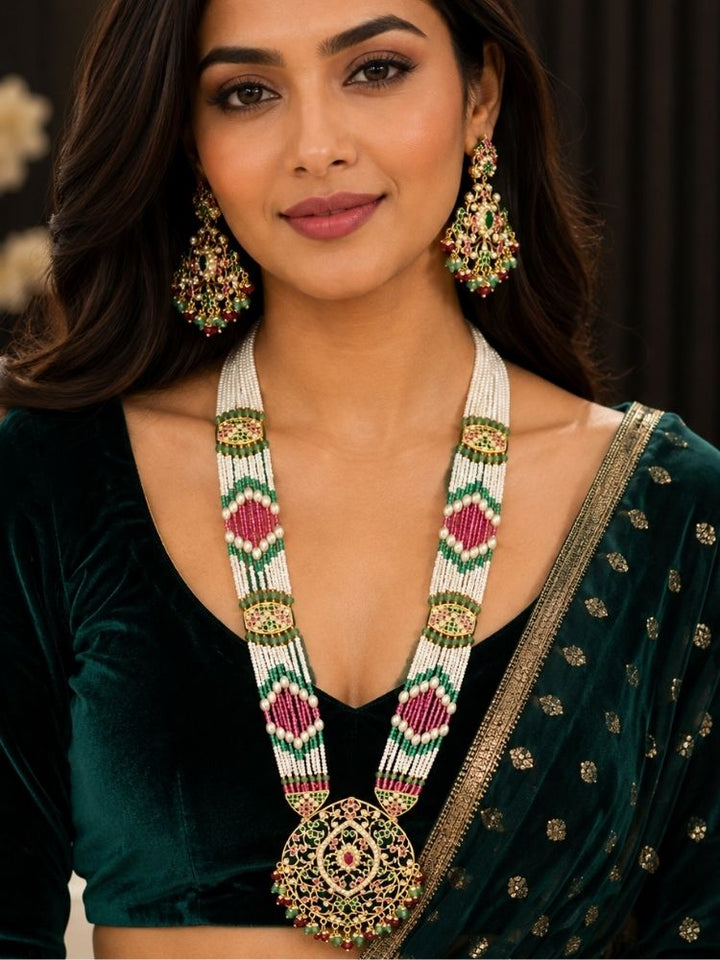 A woman wearing an ornate Indian-style jewelry set featuring a long jadau necklace with a large circular jadau pendant and matching dangling long jadau earrings. The jewelry has white beads with green and pink geometric patterns and gold-toned metal detailing.
