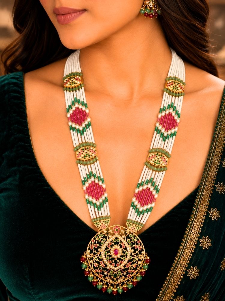 A close-up of a woman wearing a traditional Indian jadau necklace with a large ornate gold pendant decorated with red and green stones, paired with a dark green velvet top.