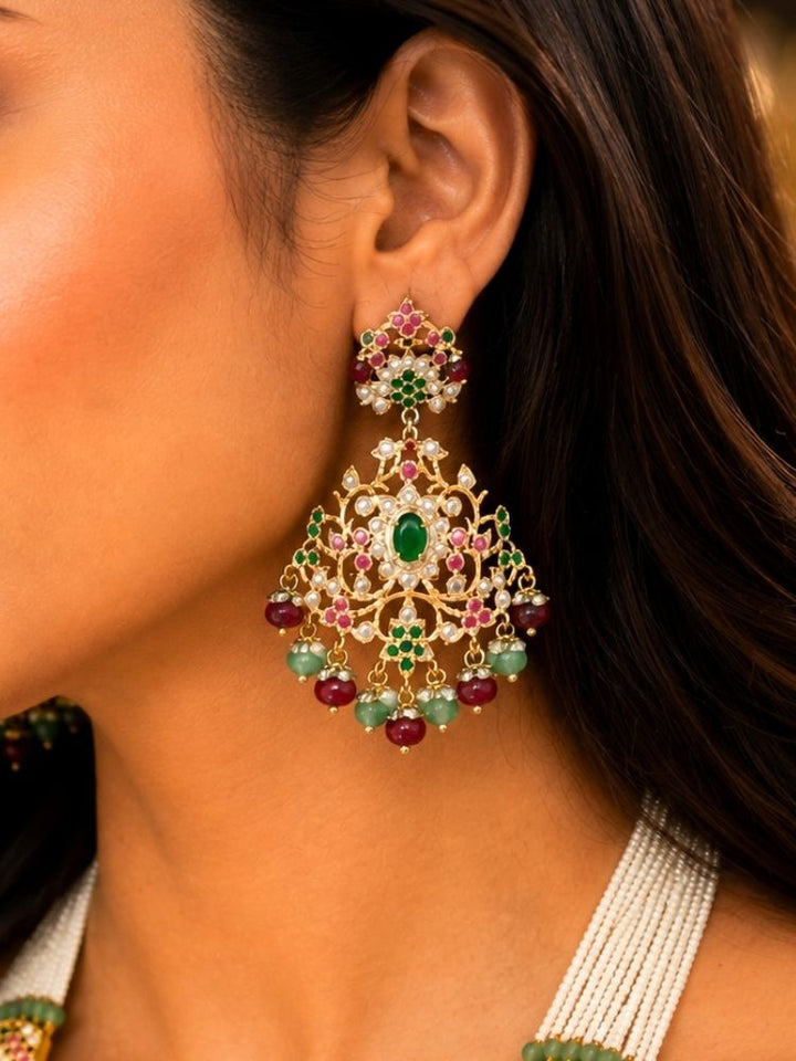 A close-up of a woman wearing a large, ornate gold-toned chandelier jadau earring decorated with green, pink, and red gemstones.