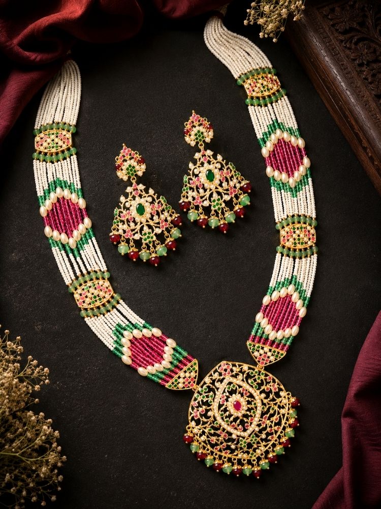 An ornate Indian jadau jewelry set featuring a jadau necklace and matching earrings, displayed on a dark textured background with draped maroon fabric and dried floral accents. The jewelry features white beads, gold-plated filigree, and vibrant pink and green beadwork.