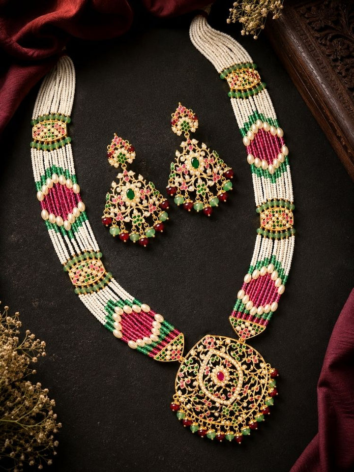 An ornate Indian jadau jewelry set featuring a jadau necklace and matching earrings, displayed on a dark textured background with draped maroon fabric and dried floral accents. The jewelry features white beads, gold-plated filigree, and vibrant pink and green beadwork.