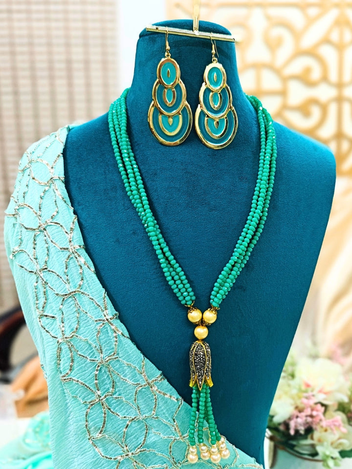 A mannequin showcases the Jade Sea Green Necklace Set—featuring a layered sea green necklace with crystal beads and gold accents, plus matching turquoise and gold dangle earrings—against a soft floral backdrop.