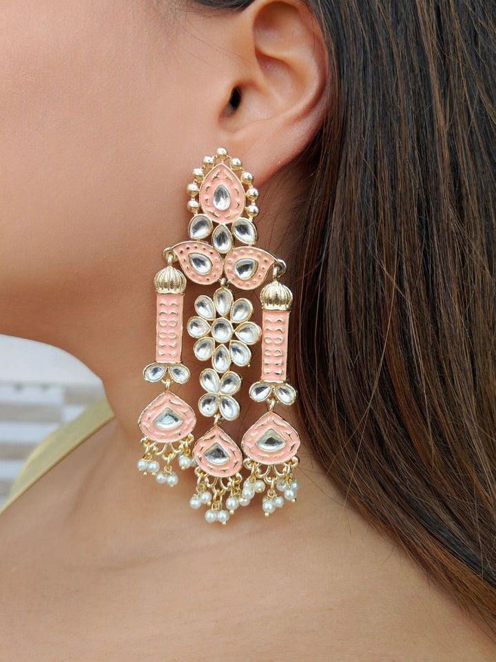 A close-up of a womans ear wearing Jasmine Peach Earrings—large, ornate Meenakari long earrings with pink and gold accents, white kundan stones, pearl bead hangings, and intricate floral and geometric patterns.